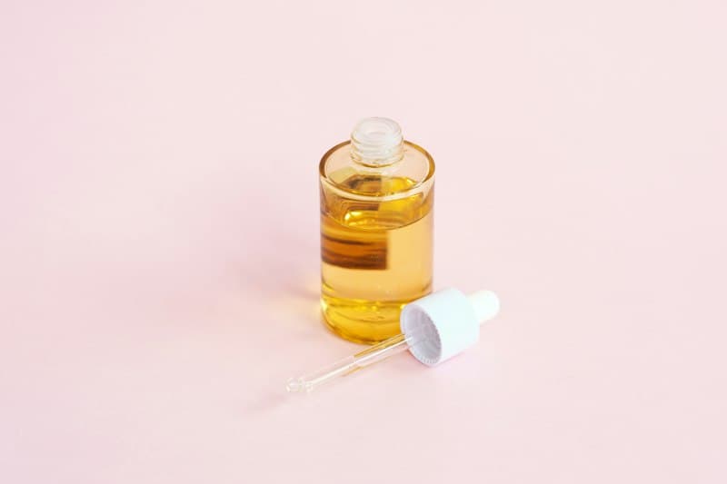 A glass bottle of golden serum with its dropper on a soft pink surface
