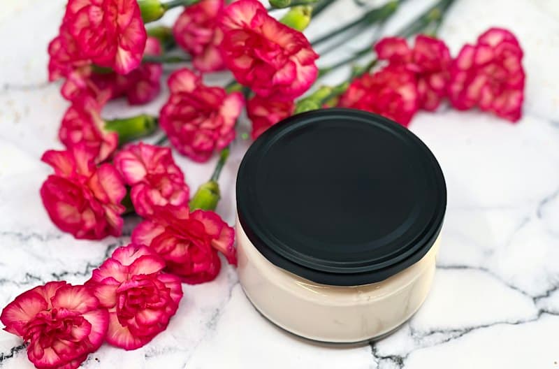 A jar of body butter with a black lid beside pink carnations on marble